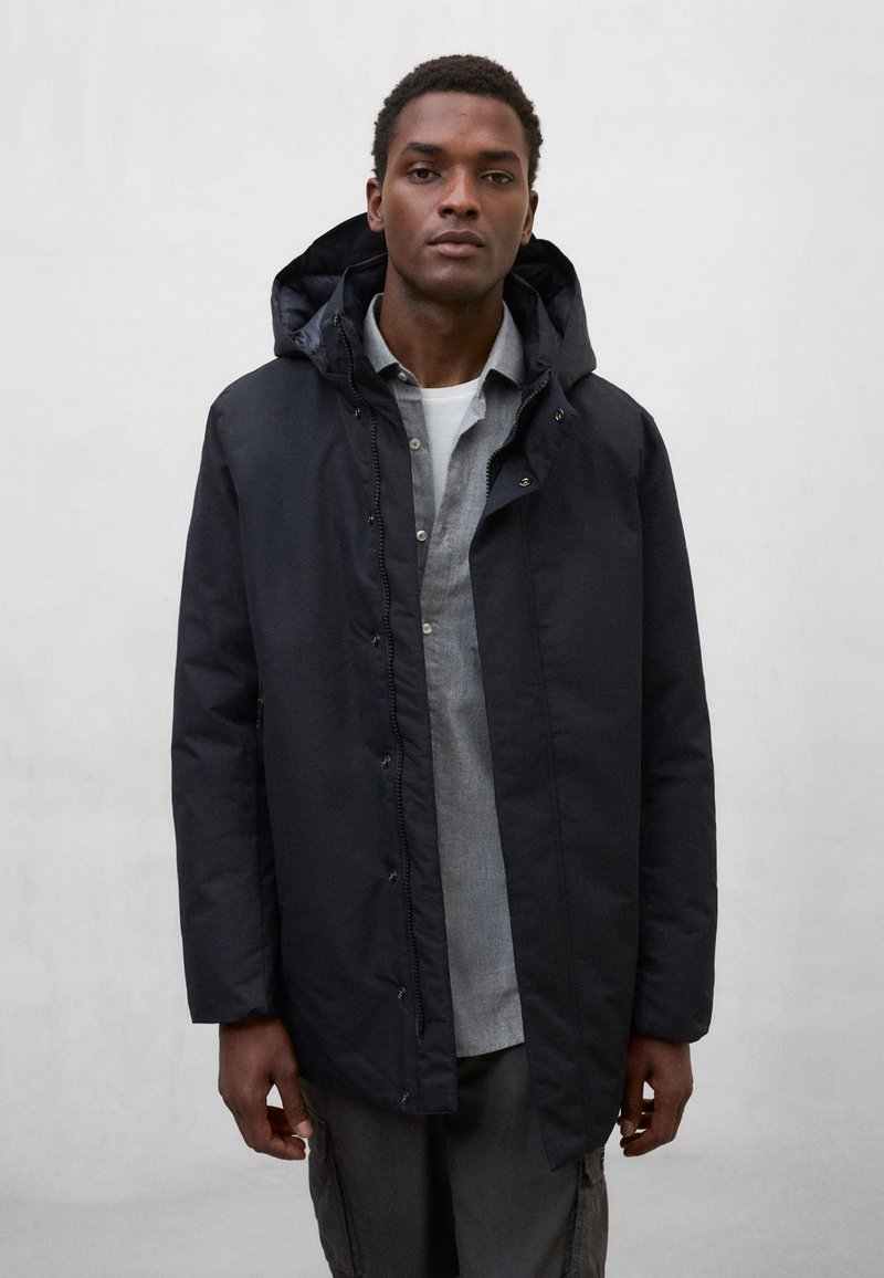 Czarna, wodoodporna parka z kapturem, wyposażona w przedni zamek błyskawiczny i zatrzaski, noszona na jaśniejszej, szarej koszuli, na jednolitym tle.