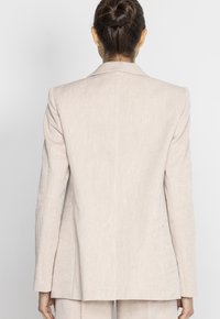 Jasnobeżowa lniana marynarka z klapami typu notch, wyraźnie zarysowanymi ramionami i prostym krojem. Widok z tyłu ukazuje czyste linie i minimalną ilość detali.