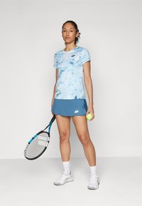 Lyseblå idrettsdrakt med et mønster, kombinert med et blått skjørt. Modellen holder en tennisracket og en gul tennisball. Hvite sko og sokker.