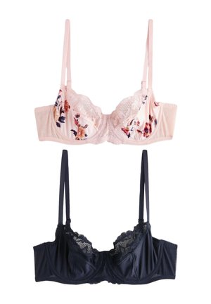 Femme portant un soutien-gorge en dentelle florale rose clair et une culotte assortie, debout contre un mur clair uni, regardant légèrement vers la droite.