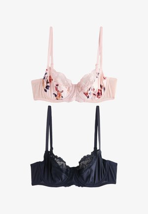 Femme portant un soutien-gorge en dentelle florale rose clair et une culotte assortie, debout contre un mur clair uni, regardant légèrement vers la droite.
