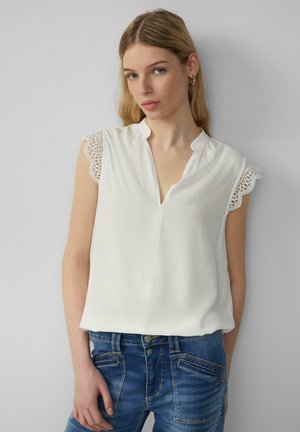 Blouse blanche avec un décolleté en V et des détails en dentelle sur les manches. Le tissu est lisse et le design est décontracté, associé à un jean en denim bleu.