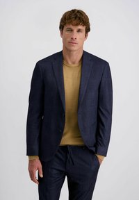 Mann mit kurzen, lockigen braunen Haaren, der einen dunkelblauen karierten Anzug und einen senfgelben Pullover trägt, steht mit einer Hand in der Tasche.