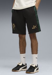 Svarta shorts med gröna mönstrade sidostripes, gyllene "KING"-tryck och en diamantlogotyp. Bärs med svarta skor och vita strumpor.