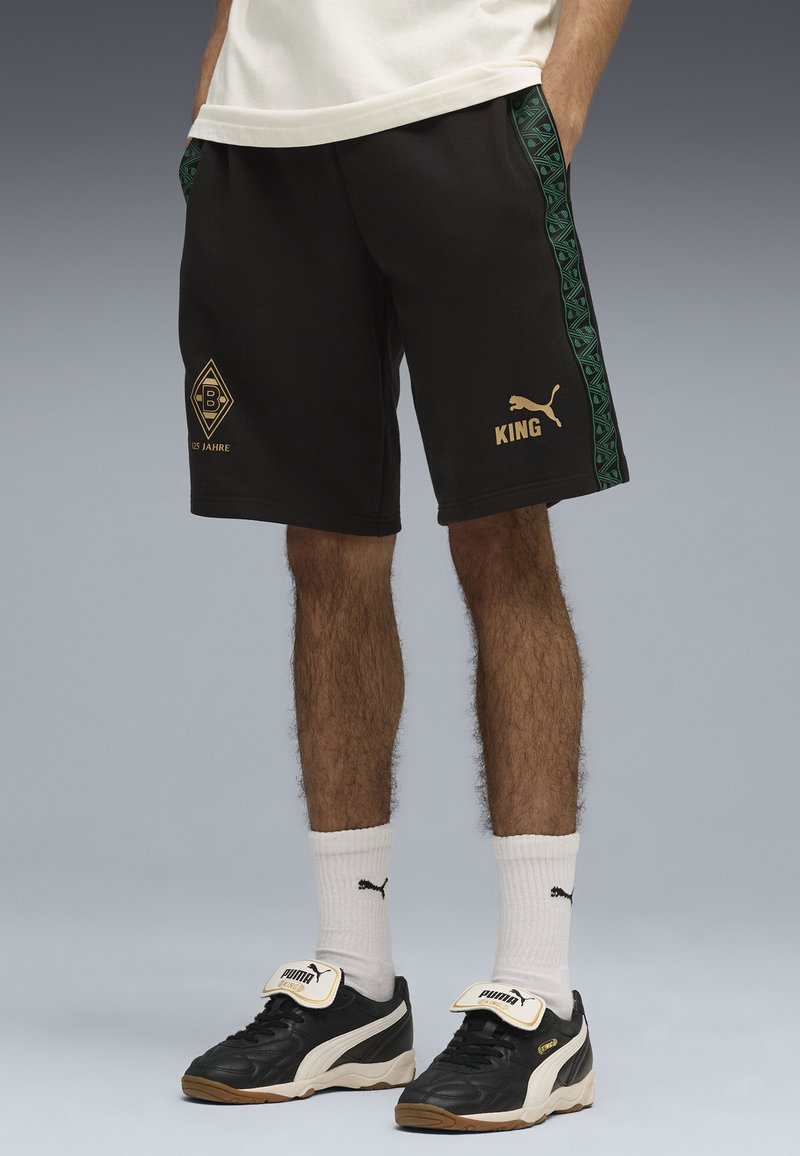 Svarta shorts med gröna mönstrade sidostripes, gyllene "KING"-tryck och en diamantlogotyp. Bärs med svarta skor och vita strumpor.