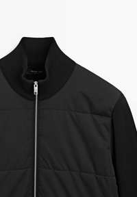 Giacca nera trapuntata con collo alto a coste e chiusura frontale con zip. Realizzata con materiali a contrasto, texture liscia e design semplice.