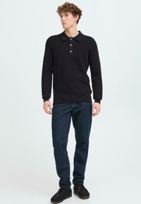 Fekete kötött pulóver galléros nyakkal és háromgombos patenttal, sötétkék farmernadrággal és fekete cipővel párosítva.