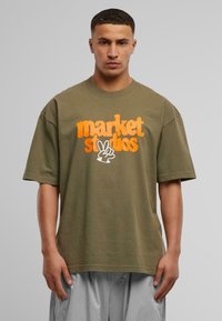 Olivenfarget oversized t-skjorte laget av bomull med oransje tekst "market studios" og en hvit håndgrafikk. Korte, brede ermer.