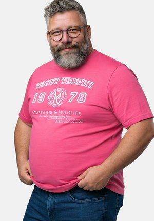 Uśmiechnięty, brodaty mężczyzna w okularach, ubrany w różowy t-shirt z napisem "Trout Trophy 1978 Outdoor & Wildlife" oraz niebieskie jeansy, trzymający za rąbek koszulki.