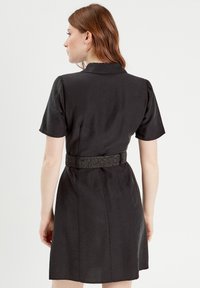 Robe noire courte avec des manches courtes, un col structuré et une ceinture texturée. Le tissu semble lisse avec un léger éclat.