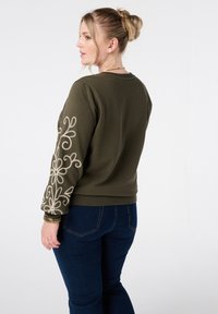 Olijfgroene sweatvest met decoratieve witte bloemenborduursels op de mouwen, zachte stof, geribbelde boorden en zoom, gecombineerd met donkerblauwe jeans.
