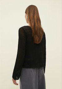 Pull noir en tricot ajouré avec manches longues et coupe décontractée. Motif texturé avec ourlet légèrement irrégulier. Porté sur un pantalon gris ample.
