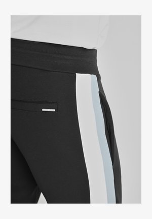Pantalones de chándal negros con paneles laterales en blanco y azul claro, con una pretina texturizada y un pequeño logotipo metálico en el bolsillo.