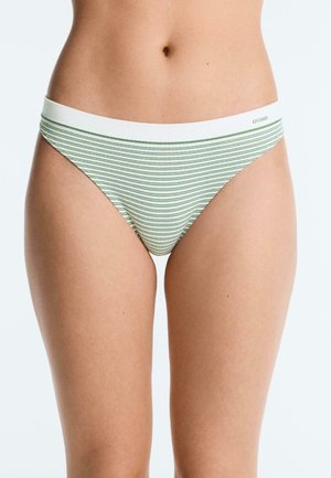 Femeie purtând lenjerie de baie de tip bikini albă, cu dungi orizontale subțiri verzi și o bandă albă în talie cu inscripția "OYSHO".