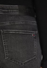 Los vaqueros de mezclilla negros presentan un bolsillo trasero con costuras blancas y una etiqueta de cuero negro texturizada, con el logotipo "TOMMY HILFIGER" en relieve.
