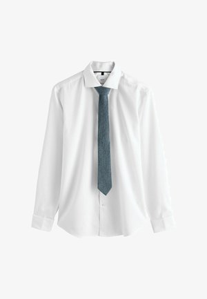 Balta ilgomis rankovėmis marškiniai su klasikinio stiliaus apykakle, derinami su tekstūruotu tamsiai mėlynu kaklaraiščiu. Marškiniai užsegti ir tvarkingai išlyginti.