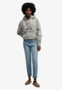 Szara bluza z kapturem z napisem „Long Island University”, zestawiona z jasnoniebieskimi dżinsami i beżowymi wsuwanymi butami. Teksturowany materiał i luźny krój.