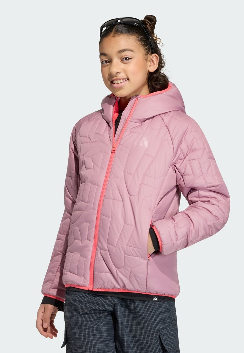 Fille souriante portant une veste matelassée rose clair avec capuche, des lunettes de soleil sur la tête, et un pantalon à carreaux foncé.