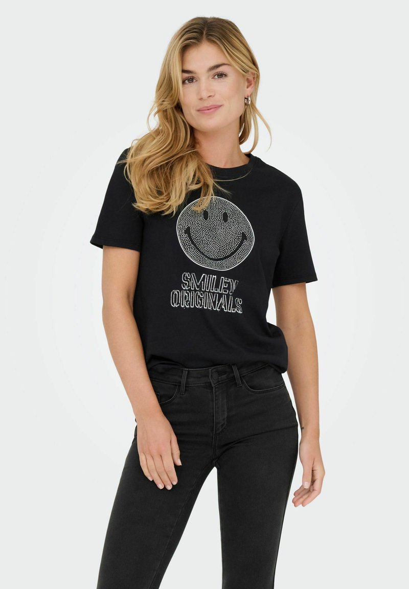 T-shirt noir avec un grand graphique de visage souriant blanc et le texte "SMILEY ORIGINALS" en dessous. Le modèle porte un jean noir taille haute.