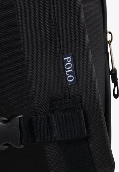 Čierny látkový ruksak s hladkou textúrou, nastaviteľný popruh s plastovou prackou a malou modrou nálepkou s textom "POLO".