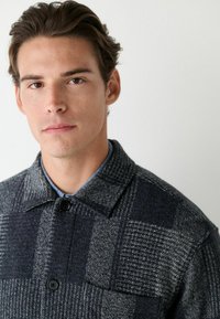 Šedý a černý kostkovaný vlněný kabát s límcem, velkými knoflíky, texturovanou tkaninou a náprsní kapsou na levé straně.