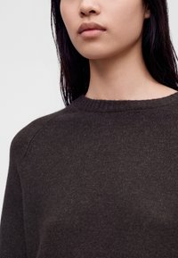 Femme aux cheveux raides et foncés portant un pull en maille marron foncé à encolure ronde sur un fond clair uni.
