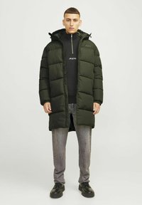Dlouhý zelený péřový kabát s kapucí, na zip a velkými prošívanými částmi. Nosený přes černou mikinu na zip a šedé džíny. Černé boty.