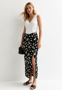 White fitted top with a V-neck, paired with a black floral maxi skirt featuring yellow daisies and a side slit. Black sandals and bag.