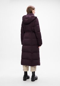 Lang, lilla pufferjakke med hætte, der har en tætsiddende talje, kviltet design og en glat tekstur. Båret over beige bukser og sorte støvler.