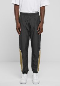 Sorte træningsbukser med elastisk talje, med guld sidepaneler og hvid piping. Stylet med hvide sneakers. Glat, letvægtsstof.
