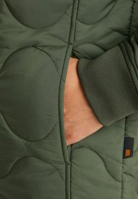 Veste matelassée verte avec une poche latérale, des poignets côtelés et des motifs de couture visibles. La texture semble lisse et isolante.