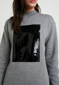 Sudadera gris con un cuello alto que presenta un rectángulo negro brillante impreso con "CALVIN JEANS" en letras negritas.