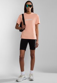 Pfirsichfarbene Baumwoll-T-Shirt mit Logo, schwarze dehnbare Shorts mit Seitenbranding, weiße Socken und Sneakers. Zubehör umfasst reflektierende Sonnenbrillen.