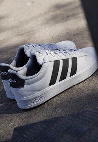 Baskets blanches Adidas avec trois bandes noires sur le côté, placées sur une surface grise à la lumière naturelle.