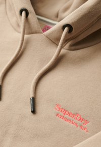 Hoodie beige con cordini, caratterizzato da un logo ricamato rosa a contrasto. Tessuto morbido con dettagli in hardware nero.