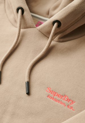 Beige hoodie met koordjes, met een contrasterend roze geborduurd logo. Zachte stoftextuur met zwarte hardware accenten.