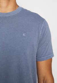 T-shirt en coton bleu avec col rond, manches courtes et un logo cousu "AE" sur le côté gauche de la poitrine. Texture lisse avec une coupe décontractée.