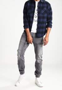 Camicia di flanella a quadri blu navy, t-shirt bianca con grafica sotto, pantaloni joggers grigi con tasche e sneakers bianche. Outfit casual.