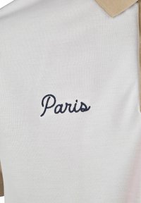 Polo blanc avec un col beige. Présente le texte brodé "Paris" en cursif marine. Fabriqué en matériau texturé.