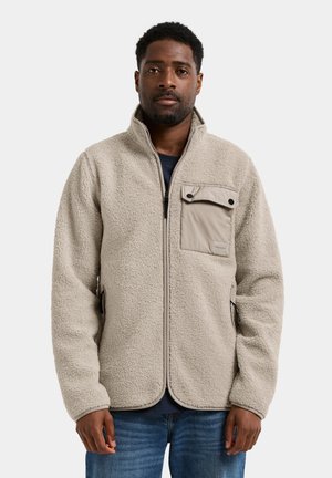 Mann trägt eine beige Fleecejacke mit Reißverschlusstaschen und einer mit Knöpfen verschlossenen Brusttasche, steht vor einem einfarbigen hellen Hintergrund.