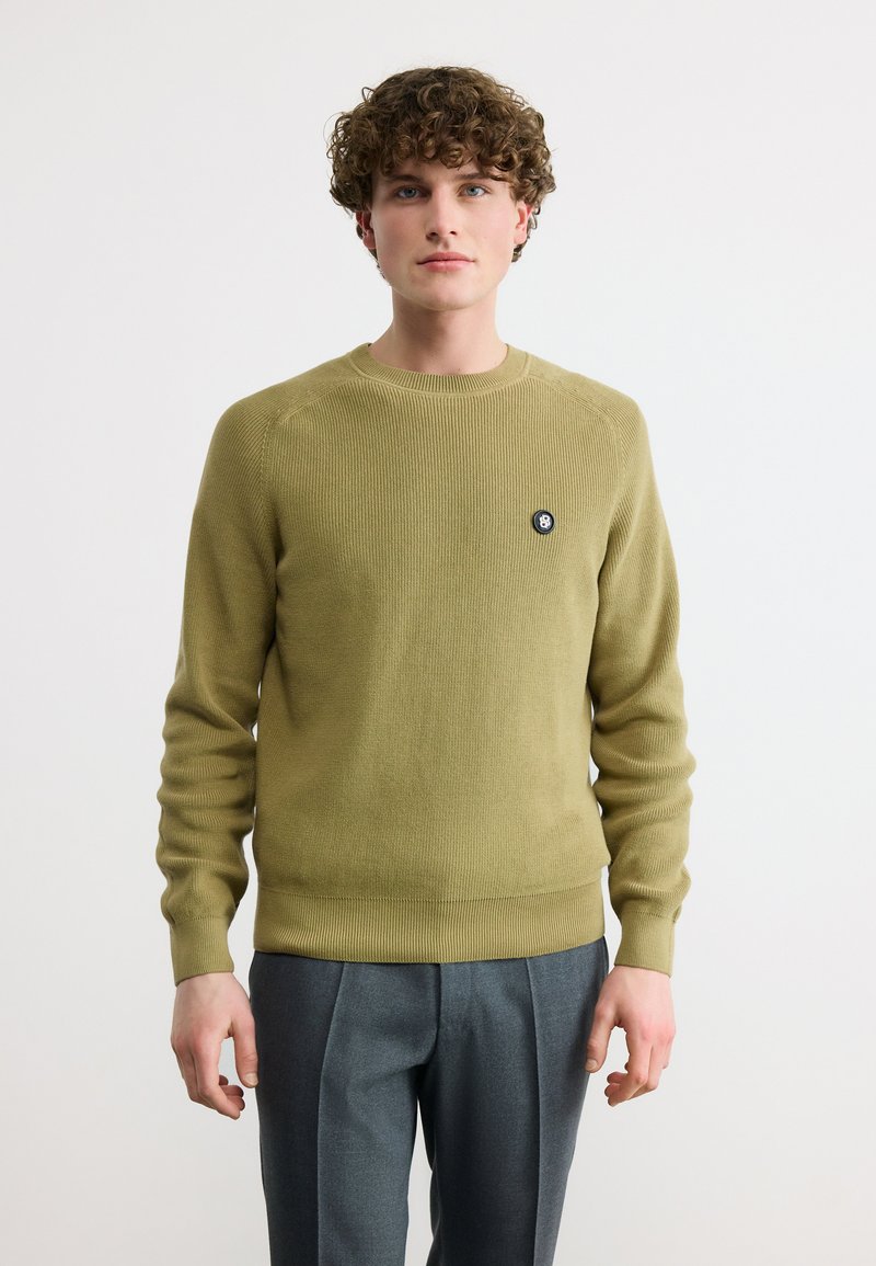 Νεαρός άνδρας με σγουρά μαλλιά, φορώντας πουλόβερ ελιάς και γκρι παντελόνι, στέκεται μπροστά σε απλό φωτεινό φόντο.