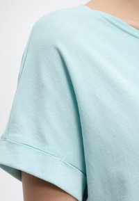 Ανοιχτό μπλε κοντομάνικο πουκάμισο με διπλωμένο άκρο στο πάνω μέρος του βραχίονα, φορεμένο από ένα άτομο με φόντο ένα απλό ανοιχτόχρωμο φόντο.