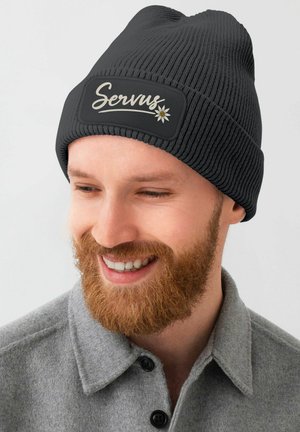 Schwarze gerippte Strickmütze mit einem rechteckigen Patch, auf dem das Wort „Servus“ und eine Blumengrafik zu sehen sind. Weicher Stoff, enganliegendes Design.