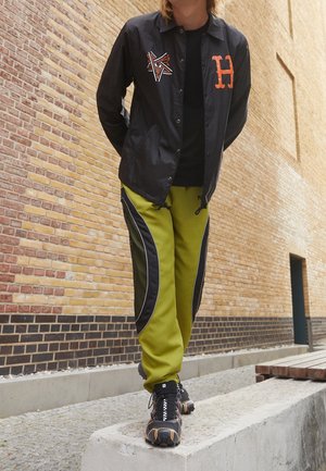 Person trägt eine schwarze Jacke mit orangefarbenen Logos, grüne und schwarze Hosen sowie schwarze Sportschuhe und steht auf einem Betonsims vor einer Backsteinmauer.