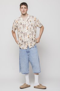 Rövid ujjú bézs ing színes kéz- és edénymintákkal, kék, oversize farmernadrággal párosítva, fehér zoknikkal és barna klumpákkal.