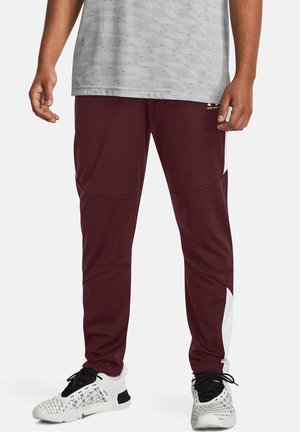 Man draagt bordeauxrode sportbroek met witte zijpanelen en witte sneakers met zwarte veters, staand tegen een effen achtergrond.
