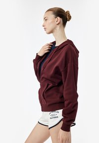 Sudadera con cremallera color burdeos, con capucha forrada, puños de canalé y bolsillos delanteros, llevada sobre una camiseta azul marino, combinada con pantalones cortos blancos con acentos en azul marino.