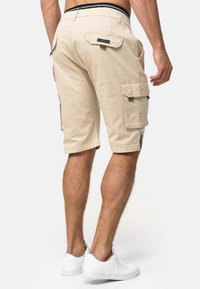 Shorts cargo beiges avec plusieurs poches latérales, un tissu texturé et une ceinture noire. Portés avec des baskets blanches.