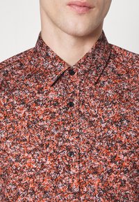 Camicia a maniche corte con un motivo rosso e nero, con colletto e chiusura con bottoni; il tessuto ha un aspetto strutturato.