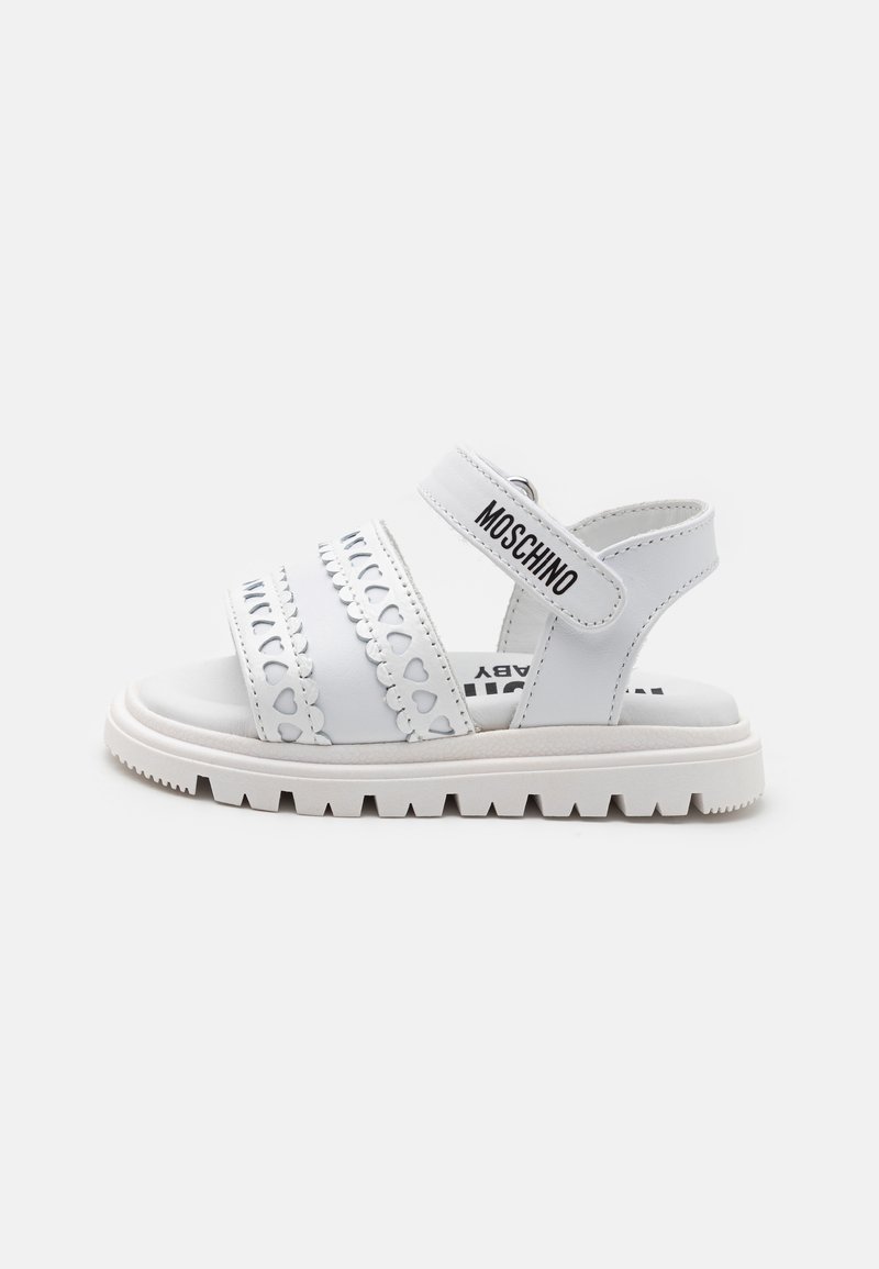White leather sandal with a thick sole, featuring cut-out heart designs and adjustable straps. Branding visible on the strap.